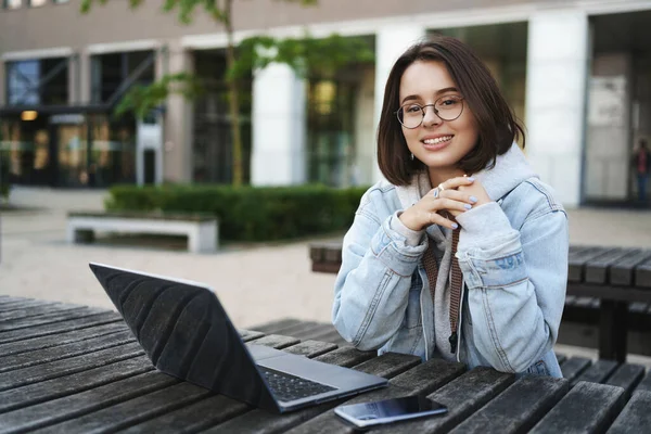 Freelance, mensen en onderwijs concept. Vrolijk jong aantrekkelijk meisje zit alleen op park bank, universiteit, werken op afstand met laptop, mobiele telefoon, weg te kijken met een tevreden glimlach — Stockfoto