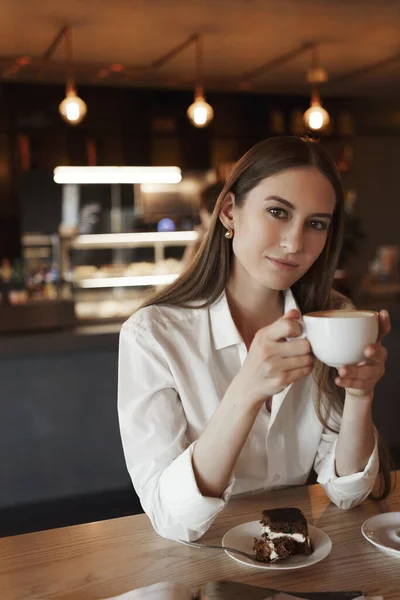 Potret vertikal wanita muda romantis yang minum kopi sendirian di kafe yang nyaman, bersandar di meja kayu, memegang cangkir dan tersenyum di depan kamera, makan kue saat makan siang Stok Gambar