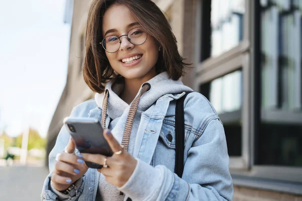 Potret close-up dari perempuan muda yang menarik dengan jaket denim, menunggu pasangannya, menggunakan aplikasi ponsel, melihat kamera dengan senyum berseri-seri, berdiri di jalan di pagi hari Stok Gambar