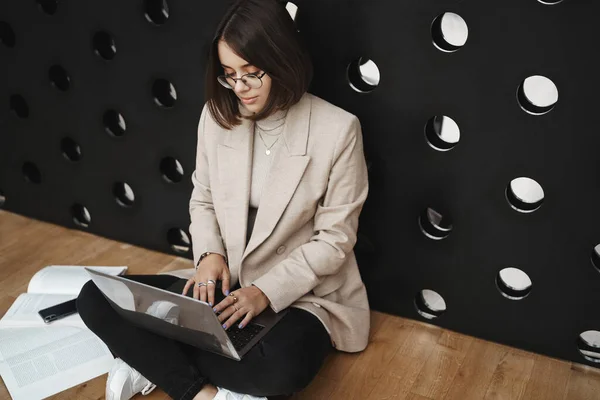 Close-up portrait of smart cute young woman sit on floor and work on project, writing in laptop, look computer screen studying homework, prepare work report or presentation, lean wall Stock Image