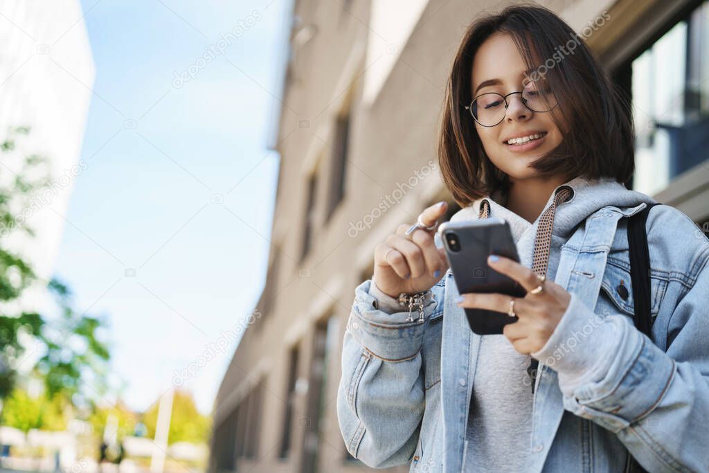 Low angle outdoor portrait of smiling pretty girl using mobile phone, smiling at smartphone display, use app to order taxi, messaging on her way to party, stand on street in glasses and denim jacket