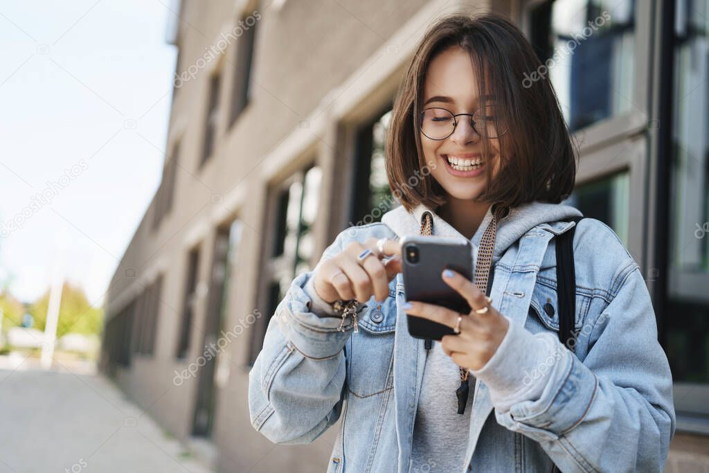 Youth, technology and spring concept. Cheerful pretty young woman in glasses, denim jacket, laughing and smiling looking mobile phone display, send funny message, texting as standing on street
