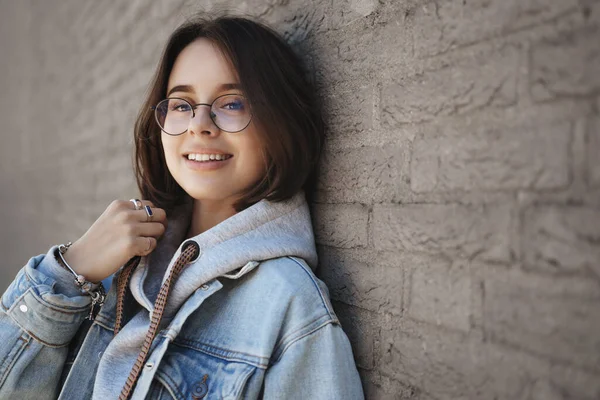 Retrato de cerca de una alegre y sonriente chica de pelo corto con gafas y chaqueta de mezclilla, apoyada en la pared del edificio, mirando a la cámara con expresión feliz y despreocupada, disfrutando de caminar afuera. —  Fotos de Stock