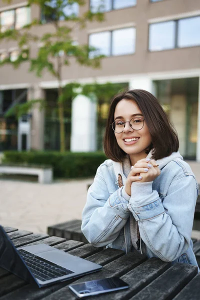 Verticaal schot van glimlachende vrolijke queer meisje zitten in het park op bank, werken ver van buiten, met behulp van laptop, freelancer doet haar werk, lachen genieten van frisse lente lucht — Stockfoto