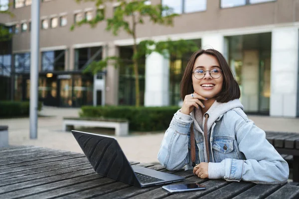 Portret van gelukkig modern meisje buiten zittend met laptop op tafel, wegkijkend en lachend vrolijk als werkend weg van kantoor, afstandsonderwijs, wachtend op iemand in het park — Stockfoto