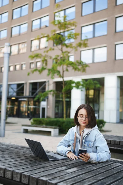 Verticaal portret van jonge leuke vrouwelijke student, meisje zit buiten met laptop, kijken naar mobiele scherm tijdens het werk, smm antwoord op vragen van klanten, studeren online — Stockfoto