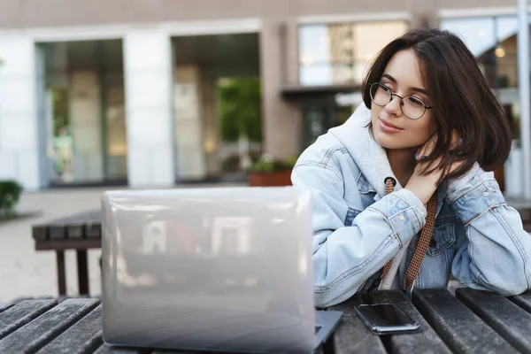 Potret close-up dari gadis muda yang cantik dalam kacamata dan jaket denim, bersandar di atas meja dengan wajah santai bijaksana, telah istirahat dari belajar, menggunakan proyek laptop menulis atau bekerja lepas — Stok Foto