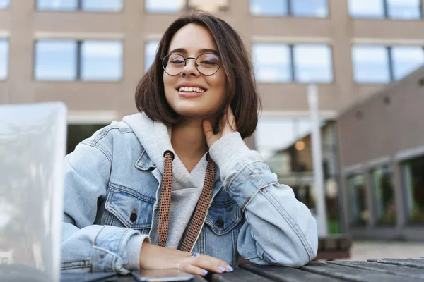 Close-up portrait of young attractive queer girl in denim jacket, student sitting outside with laptop, smiling at camera with pleased calm expression, have a break from work on project — Stock Photo, Image