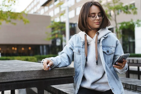 Portrait of young modern hipster girl in denim jacket, glasses sitting outdoors at city square, park bench, turn aside to check messages on mobile phone, smiling as texting friend, use application — Stock Photo, Image