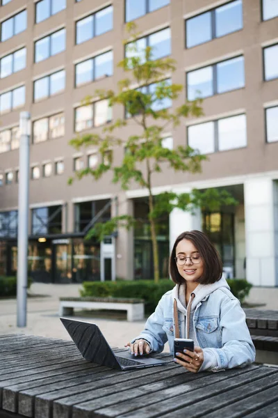 Ritratto verticale di giovane donna carina sorridente seduta all'aperto circondata da edifici, utilizzando il telefono cellulare e il computer portatile, sorridendo al display dello smartphone, lavorando a distanza, freelance Immagine Stock