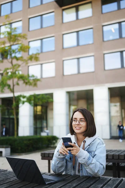 Vertical portrait of young woman sitting alone outdoors, looking away with dreamy happy smile, texting on mobile phone, have break from studying, writing report laptop, working on remote Royalty Free Stock Photos