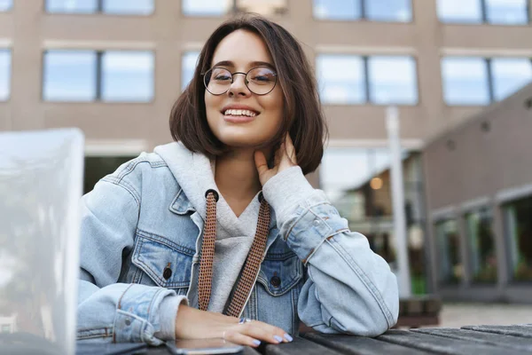 Ritratto ravvicinato di giovane attraente ragazza queer in giacca di denim, studente seduto fuori con il computer portatile, sorridente alla fotocamera con espressione calma piacere, hanno una pausa dal lavoro sul progetto Foto Stock Royalty Free