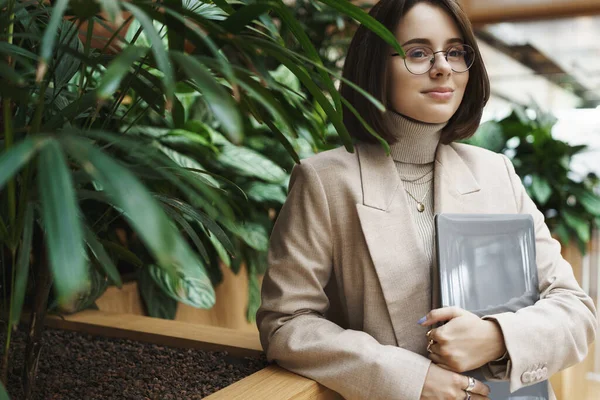 Career, work and women concept. Close-up portrait of successful young woman in beige jacket waiting for coworker at reception hotel or office building, hold laptop, smiling and watching passersby Royalty Free Stock Images