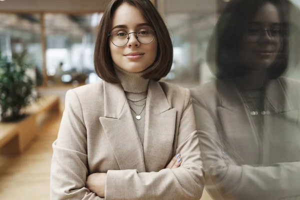 Portrait of confident good-looking businesswoman, girl in blazer and glasses, apply for job in IT company, lean on wall in office hall, lobby, cross hands chest self-assured smiling — Stock Photo, Image