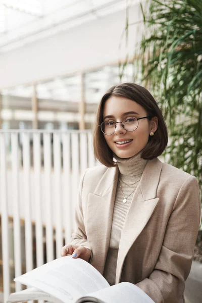 Vertical portrait of attractive elegant young woman in glasses and blazer, sitting in hall, business center, teaching foreign language, prepare homework or material for next classes to teach — Stock Photo, Image