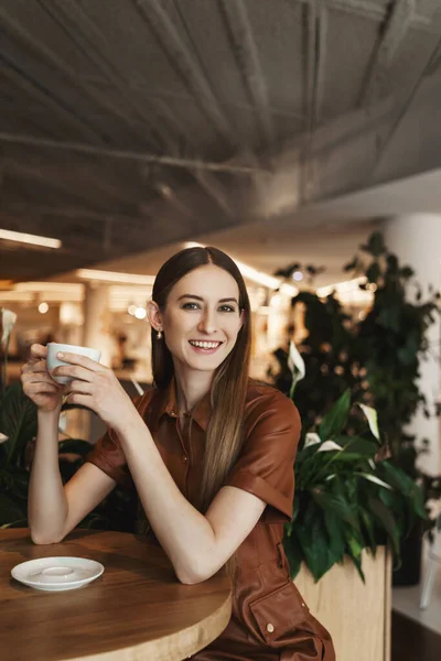 Retrato vertical de una mujer joven y elegante disfrutando de una buena taza de café, sentado en la cafetería, teniendo una conversación optimista con su novia, sonriendo ampliamente cámara, viejos amigos se reúnen para chatear —  Fotos de Stock