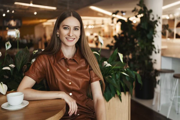 Negocios, concepto de carrera femenina. Retrato de cerca de la elegante encantadora joven que se inclina mesa de café en la cafetería, beber capuchino o té, sonriendo con alegría cámara, gerente de restaurante en el descanso — Foto de Stock