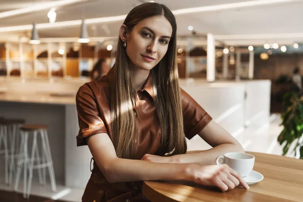 Concepto de mujeres empoderadas. Atractiva joven empresaria exitosa sentada en la cafetería, bebiendo café taza, cabeza inclinada y mirando la cámara con interés, sonriendo seductora — Foto de Stock