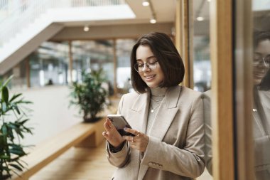 Şirket için çalışan başarılı genç bir kadının portresi, koridorda iş arkadaşını beklemek, cep telefonundan mesajları kontrol etmek, akıllı telefon kullanmak ve mutlu gülümsemek.