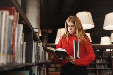 Kütüphanede rafların yanında duran, kitap okuyan, bir sonraki ders için ödev yapan, yeni şeyler öğrenen, yeni yetenekler edinen zeki kızıl saçlı kızın yan görüntüsü.