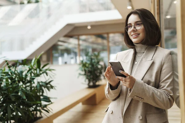 Portrait of talented, successful female employee, entrepreneur waiting for client in hall of office or business center, holding mobile phone, messaging client, smiling satisfied, look confident — Stock Photo, Image