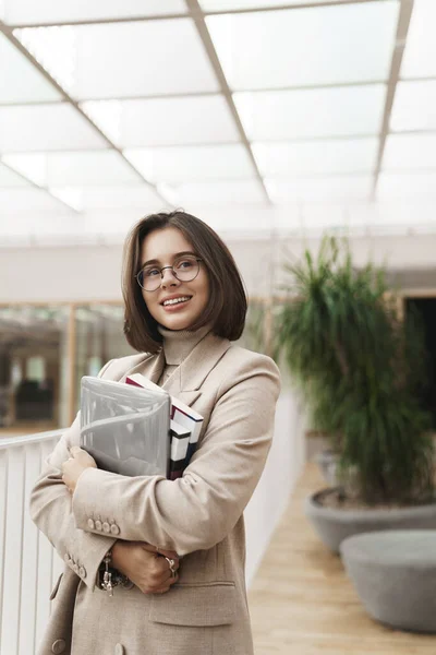 Ritratto verticale di giovane donna di successo, dipendente o imprenditore in ufficio, tutor portare laptop e quaderni, guardando lontano con soddisfazione, sorriso soddisfatto, come il suo lavoro — Foto Stock