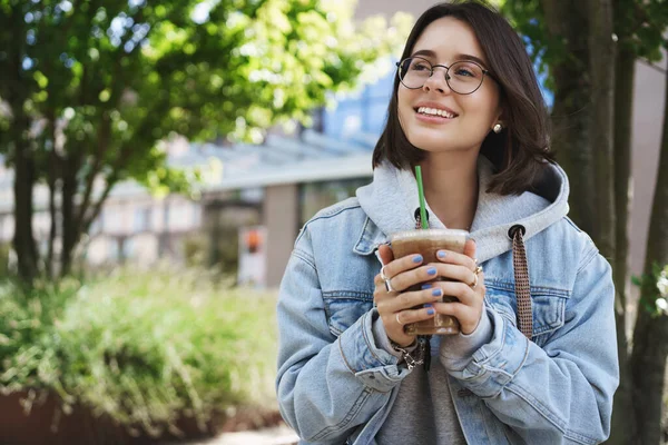 Portrait of young romantic and dreamy girl in glasses and denim jacket, looking up contemplating birds singing on bright perfect day, holding ice latte, drinking coffee and sit park bench smiling — Stok Foto
