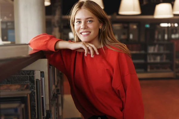 Concepto de juventud, educación y aprendizaje. Bastante sonriente pelirroja estudiante, chica se inclinan en la estantería y mirar la cámara complacido, como estudiar en la biblioteca, ratón de biblioteca recoger nueva novela para leer, hacer proyecto — Foto de Stock