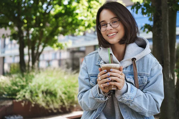 Career, women and lifestyle concept. Attractive cheerful queer girl in denim jacket, glasses, having nice conversation casually sitting park bench in spring, drinking coffee, smiling camera Royalty Free Stock Images