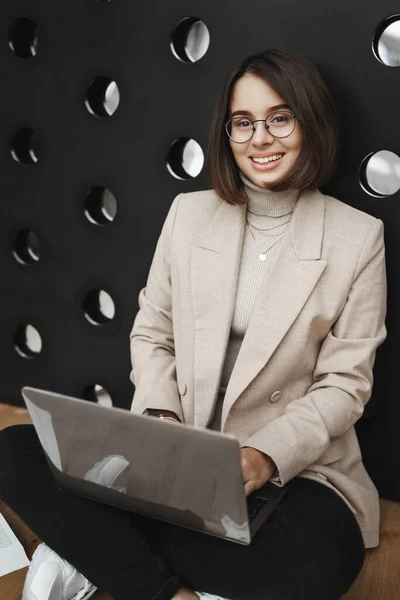 Verticaal portret van slimme stijlvolle, goed uitziende vrouw in bril en blazer, zittend op de vloer met laptop op schoot, huiswerk studeren, project voorbereiden, verslag schrijven en glimlachende camera gelukkig Stockfoto