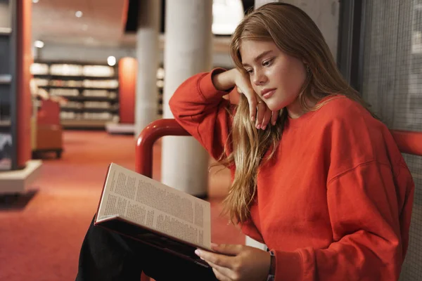 Side-view of pretty smart redhead girl reading book in library or book-crossing space, smiling tender, study in quiet place, prepared PR or research, tutor gathering info for next class — Stok Foto