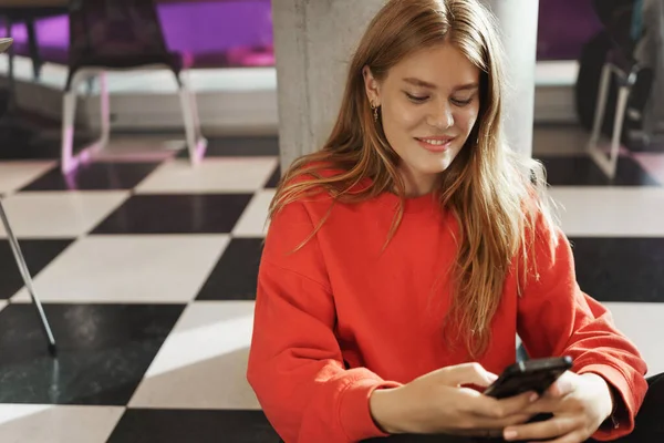 Orang muda, komunikasi online dan konsep gaya hidup. Gadis berambut merah yang mempesona duduk di lantai kafe atau ruang kerja bersama, mengirim pesan kepada teman, menggunakan ponsel dan tersenyum di layar — Stok Foto