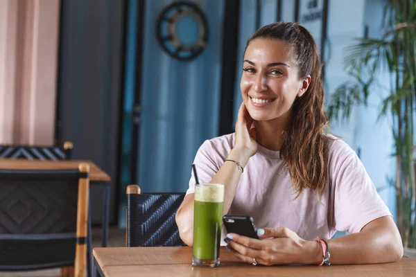 Vonzó mosolygós nő a nyaralás, élvezi paradicsom üdülőhely, ül a kávézóban a mobiltelefon és az ivás egészséges smoothie, néz kamera, önellátó időt egyedül, utazási blogger írni utáni internet — Stock Fotó