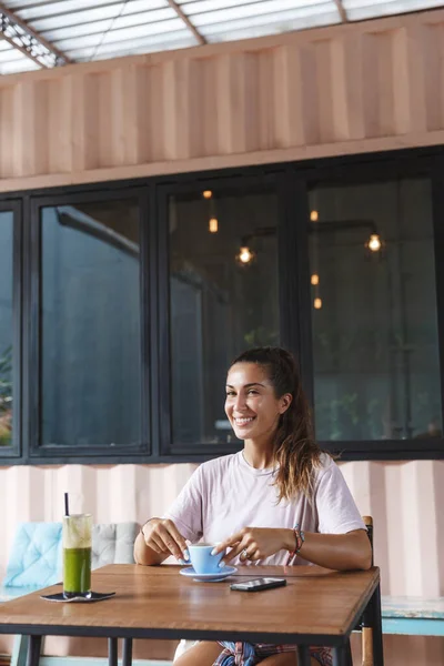 Potret vertikal wanita Eropa yang cantik duduk di meja kafe dengan smoothie dan telepon genggam, minum kopi, tertawa dan menikmati liburan yang sempurna di pulau resor Bali yang santai, tersenyum senang — Stok Foto