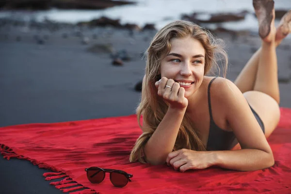 Potret wanita tropis yang bahagia dan seksi dengan pakaian renang, berbaring di handuk pantai merah di atas pantai pasir vulkanik hitam, berpaling, berjemur, menikmati liburan musim panas, menjelajahi Indonesia — Stok Foto