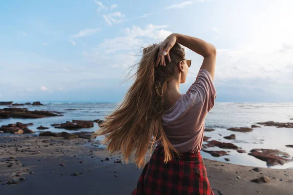 Backside view of romantic beautiful, fit hipster girl traveller long hair, enjoying blue ocean view standing black volcanic sand beach, couple trip to Bali, Indonesia, journey during summer vacation Royalty Free Stock Photos