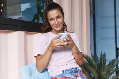 Ev molası, boş zaman ve kendini karantina konsepti. Güzel, gülümseyen bir kadın sabah çayıyla eğleniyor, kahve içiyor ve balkondan dışarı bakıyor. Covid19 salgınını izole ediyor. Mesafe koyuyor.