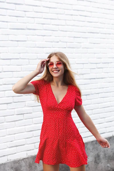 Style de vie urbain, concept estival et humain. Plan vertical de femme heureuse élégante explorant la ville, marchant sur la rue au-dessus du mur du bâtiment blanc, portant une robe rouge clair et des lunettes de soleil, souriant gai — Photo