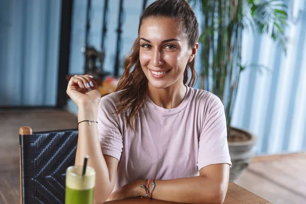 Retrato de atractiva, coqueta joven bronceada turista, chica relajante en la isla de Bali, sentarse en la cafetería de mesa de madera, bebiendo batido y disfrutando de la vista soleado complejo turístico, sonriendo feliz a la cámara —  Fotos de Stock