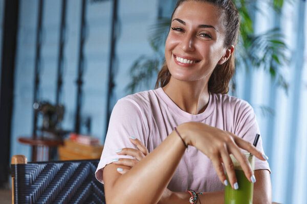 Close-up portrait of satisfied, happy relaxed female travel blogger, girl tourist sit at cafe with green smoothie, smiling camera cheerful, have vacation or honeymoon at island Bali or resort Stock Picture