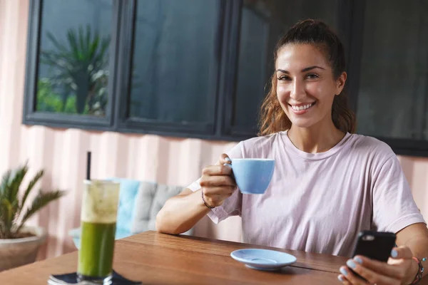 Porträt einer gesunden und glücklichen hübschen kaukasischen Frau, die mit einer Tasse leckeren Kaffees am Cafétisch sitzt, ihr Handy in der Hand hält, Nachrichten checkt, Reisebloggerin über ihren Urlaub auf den Inseln schreibt Stockbild