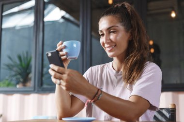 İnternet, iletişim ve tatil konsepti. Kafe masasında oturan, kahve içen, gezici ekranda gülümseyen, tatilini ve seyahat deneyimini anlatan blog yazan bir kadın portresi.