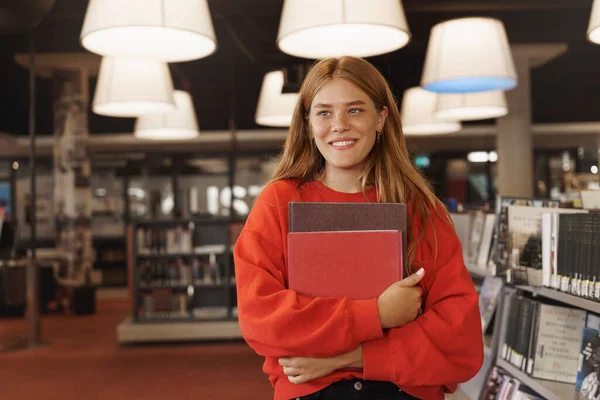 Studenti, borsa di studio e concetto di college. Bella rossa che studia, tiene libri in libreria, cerca progetto, materiale di ricerca in biblioteca, libreria dall'aspetto sorridente, gomma per esame — Foto Stock