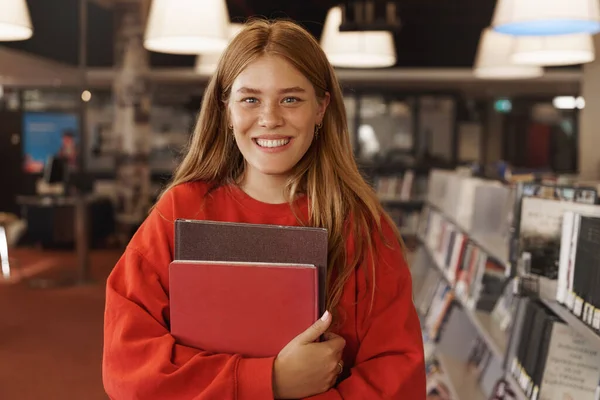 Potret siswa perempuan european berambut merah yang cantik berdiri di aula perpustakaan, toko buku retro, memegang buku dan tersenyum, belajar untuk ujian atau tes universitas, melakukan penelitian proyek untuk kursus — Stok Foto