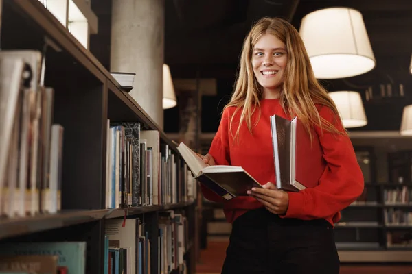 Sudut rendah ditembak cerdas gadis berambut merah yang indah membaca di perpustakaan, mengambil buku dari rak buku dan kamera tersenyum, siswa belajar, membuat penelitian untuk tes atau pemeriksaan, craming di kampus — Stok Foto