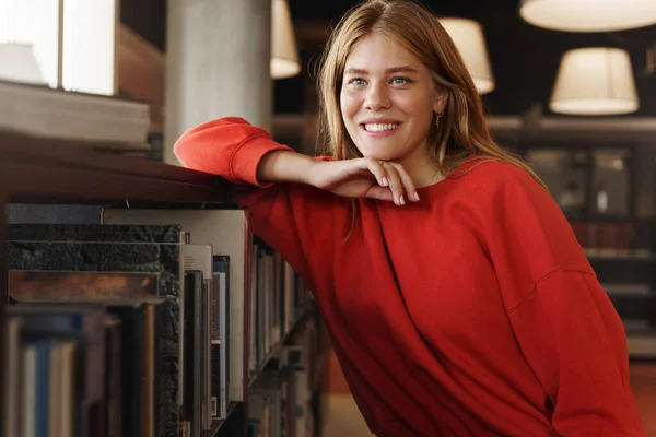 Estudiantes, carrera y concepto universitario. Mujer joven soñadora y hermosa con el pelo rojo, apoyada en la estantería, sonriendo mirando hacia otro lado en la ventana de la biblioteca, estudiando, soñando con hacerse una prueba o examen A — Foto de Stock