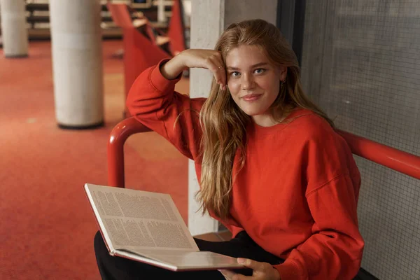 Hermosa estudiante pelirroja, chica sentada en la biblioteca con libro, leyendo para prepararse para el examen, estudiando hacer investigación, mirando la cámara con sonrisa de ensueño complacido, aprender material para las clases universitarias — Foto de Stock