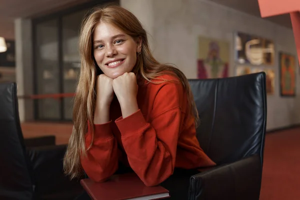 Lovely redhead female student, lean on palms and smiling as listening with interested face to friend story while sit in library, trying to study for exam, pass university test, reading books — Stock Photo, Image