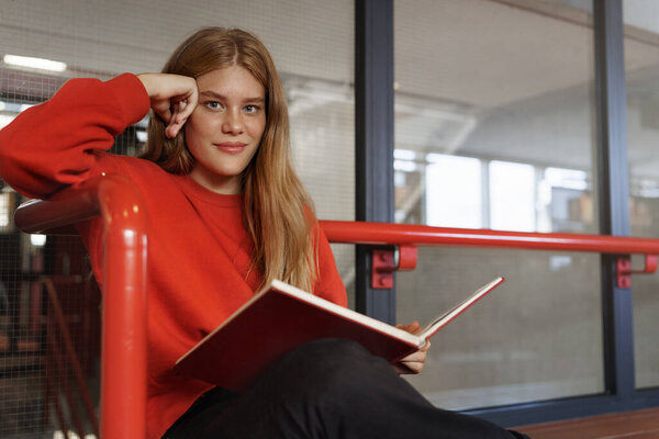 Smart attractive redhead girl, teenage student studying in library hall, reading book as sitting at campus, cramming, learning material, do research project, smile camera, bookworm likes novels Stock Photo
