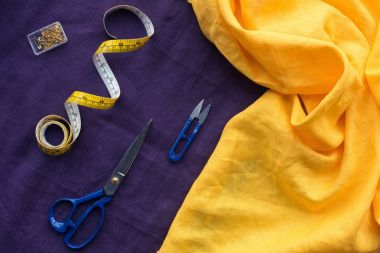 top view of scissors, measuring tape and box with pins over purple fabric